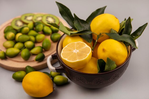 Top view of yellow lemons on a bowl with kinkans and kiwi slices on a wooden kitchen board on a white wall