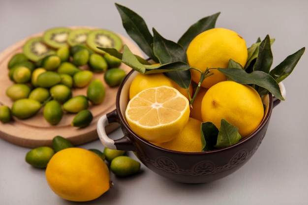 Top view of yellow lemons on a bowl with kinkans and kiwi slices on a wooden kitchen board on a white wall