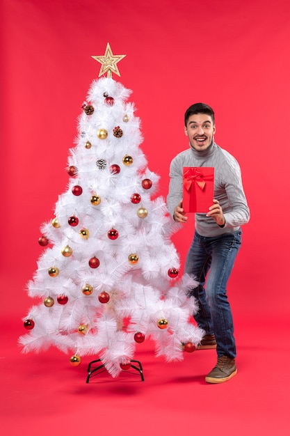 Top view of young man standing near the decorated white New Year tree and holding his gifts and showing it to his friends on red