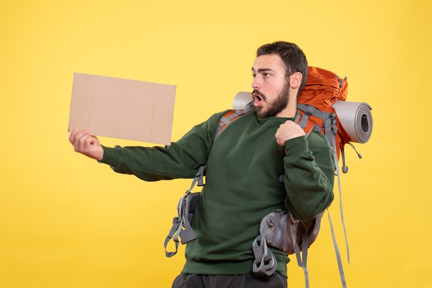 Free photo top view of young surprised and emotional travelling guy with backpack holding a sheet without writing on yellow