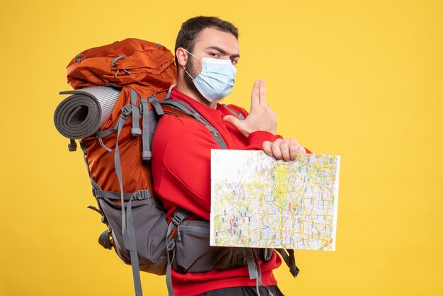 Top view of young thinking traveller guy wearing medical mask with backpack holding map pointing up on yellow