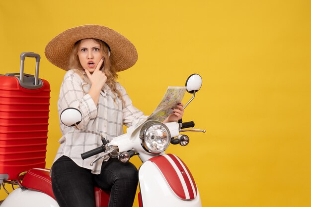 Top view of young woman wearing hat and sitting on motorcycle and holding map with confused facial epression on yellow 