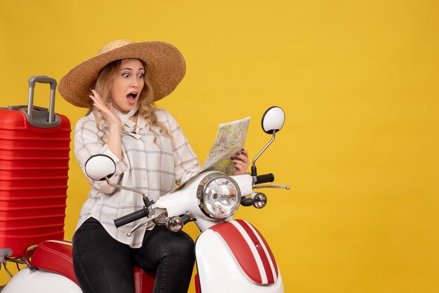 Top view of young woman wearing hat and sitting on motorcycle and looking at map feeling surprised on yellow 