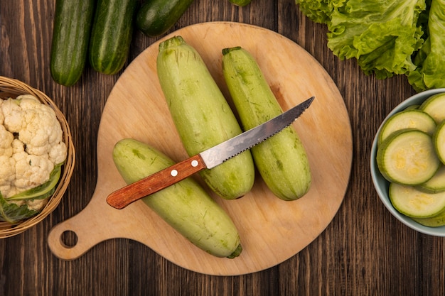 Free photo top view of zucchinis isolated on a wooden kitchen board with knife with cauliflower on a bucket with cucumbers and lettuce isolated on a wooden surface