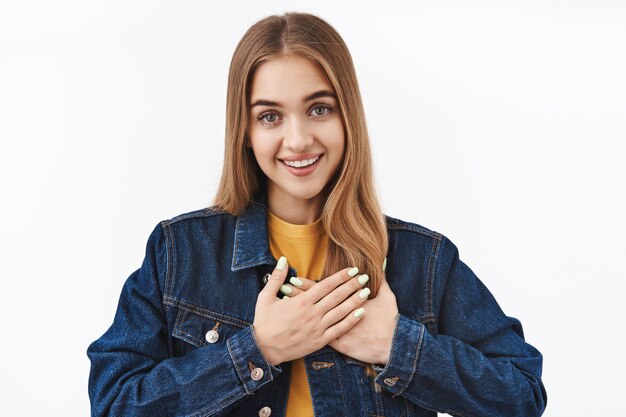 Touched happy blond girl sighing tenderly look at camera, hold hands on heart feeling grateful and impressed, standing delighted on white wall