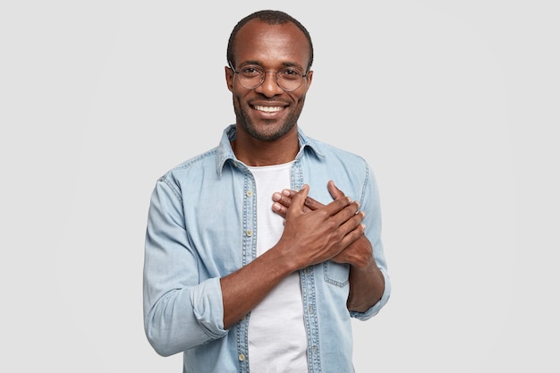 Touched and thankful African man holds hands on chest, expresses gratitude, looks pleasantly at camera