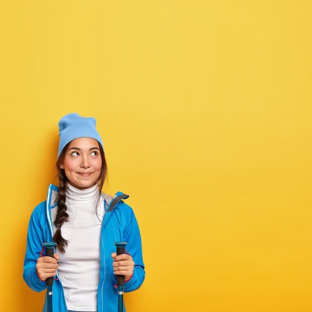 Free Photo tourism and camping concept. dreamy brunette woman uses trekking sticks, looks above, enjoys outdoor activity, looks thoughtfully above, wears blue jacket and hat, copy space on yellow wall.