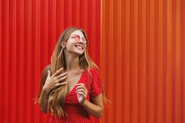 Tourism Happy woman eating sweet ice cream outdoors and looking right smiling joyful standing near red wall Female traveller exploring city on vacation