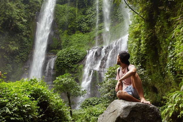 Free Photo tourism, travel and adventure. stylish young hipster sitting on stone with bare feet and turning his head back to see amazing waterfall