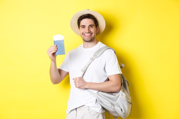 Tourism and vacation. Smiling young guy going on trip, holding backpack and showing passport with tickets, standing over yellow background