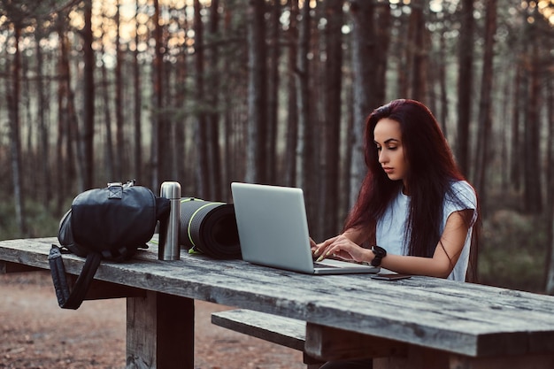 Free photo tourist hipster girl in white shirt working on a laptop while sitting on a wooden bench in the beautiful autumn forest.