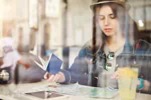 Free photo tourist sitting in cafe reading a map holding passport drinking lemonade in a busy city shot through glass