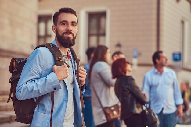 A tourist with a full beard and haircut, wearing casual clothes, holds a backpack and texting on a smartphone, standing on an antique street, during the excursion in Europe.