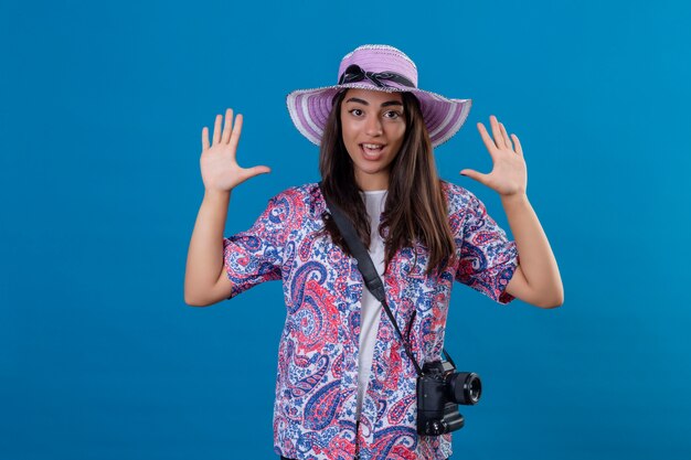 tourist woman with hat with photo camera raising hands in surrender looking surprised standing on isolated blue