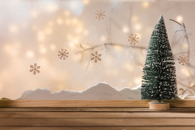 Toy fir tree on wood table near bank of snow, plant twig, snowflakes and fairy lights