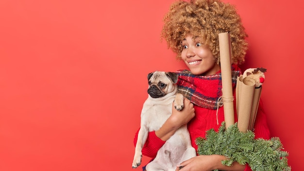 Free Photo traditional festive decoration for xmas holiday. positive curly haired woman carries rolled paper to wrap presents and green spurce wreath poses with dog indoor isolated over red background.