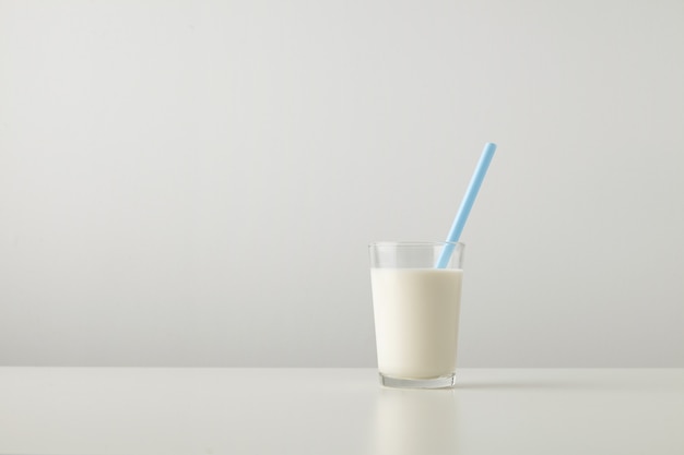 Free photo transparent glass with fresh organic milk and blue drinking straw inside isolated on side of white table