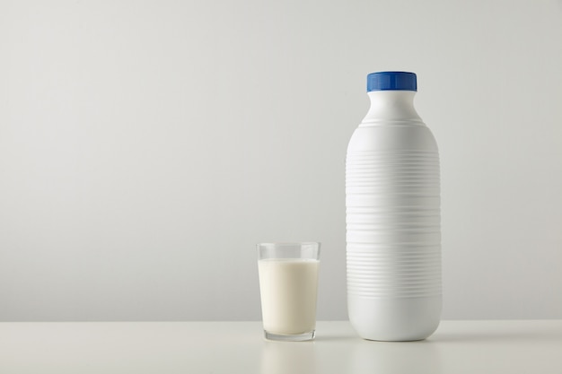 Transparent glass with fresh organic milk near plastic riffled blank bottle with blue cap isolated on side of white table.