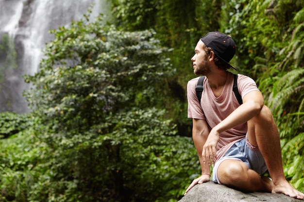 Free photo travel and adventure. handsome young barefooted male hiker with backpack relaxing alone on big stone and looking back