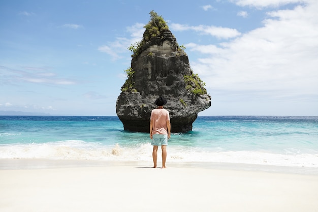 Free Photo travel, adventure and tourism. fashionable barefooted man wearing shorts, t-shirt and hat meditating at seaside, standing in front of stone island. stylish caucasian tourist admiring beautiful view