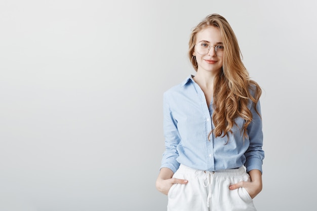 Travel agent hearing customer desires. Portrait of good-looking european businesswoman in blue blouse and glasses, holding hands in pockets and smiling, being friendly and polite over gray wall