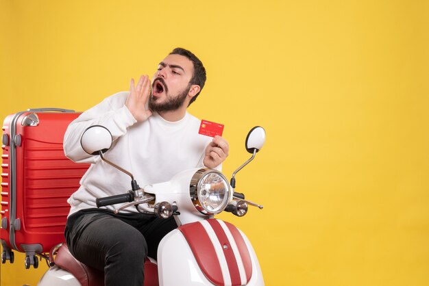 Travel concept with young travelling man sitting on motorcycle with suitcase on it holding bank card calling someone on yellow