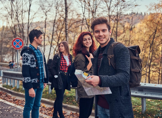 Travel, hitchhiking, adventure concept. Group of young hikers standing on the road sidelines at beautiful autumn forest, guy with map planning hike.