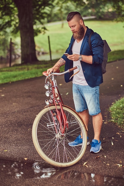 Free photo a traveler dressed in casual clothes with a backpack, using a smartphone, relaxing in a city park after riding on a retro bicycle.