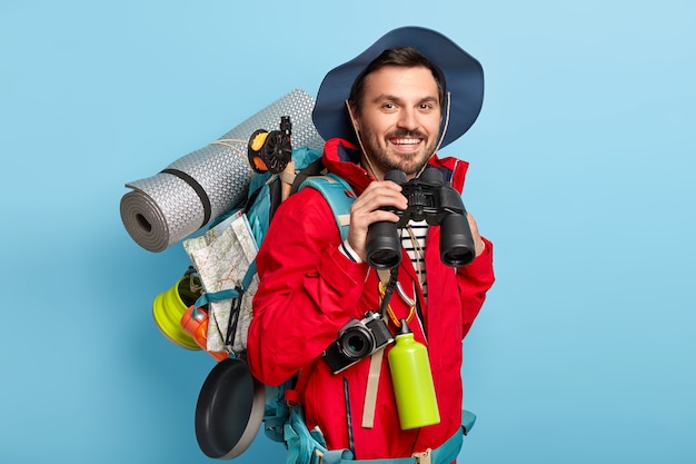 Free photo traveler man has walk with backpack, carries necessary things for trip, looks through binoculars, has glad look, wears casual clothes