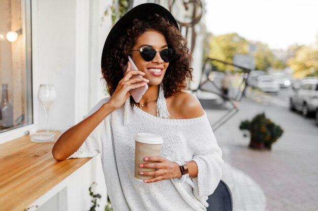 Traveling mix race  woman in stylish casual outfit relaxing outdoor  in city cafe , drinking coffee and chatting by mobile phone. Wearing trendy accessories and sunglasses.