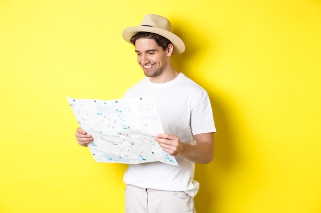 Travelling, vacation and tourism concept. Smiling happy tourist looking at map with sightseeings, standing against yellow background.