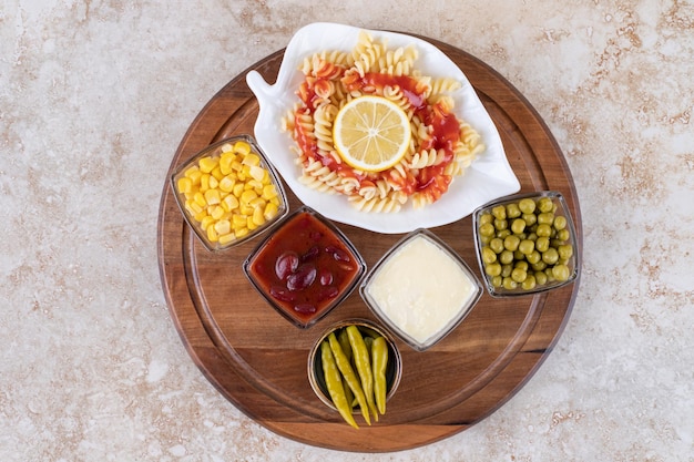 Tray with pasta serving and bowls of toppings and dressings on marble surface.