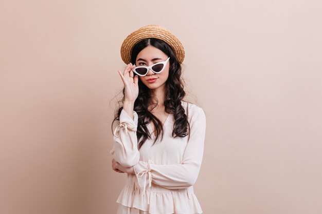 Trendy asian woman looking through sunglasses. Front view of joyful chinese lady in straw hat.