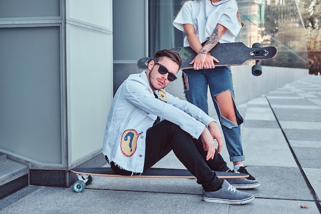 Trendy dressed couple - young hipster man sitting on longboard and his girlfriend standing near outdoors.