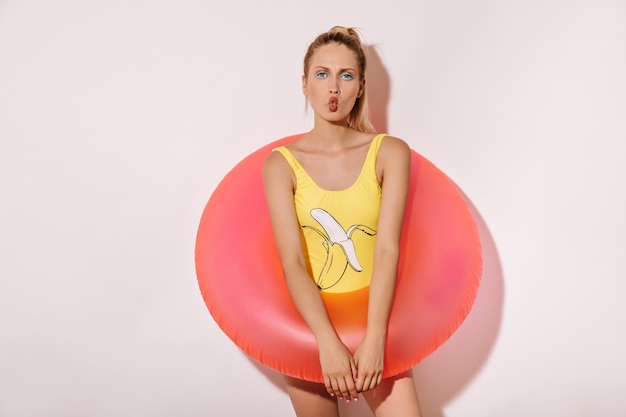 Free photo trendy girl with bright makeup in in yellow banana pattern swimsuit looking into camera and large pink swimming ring on white wall