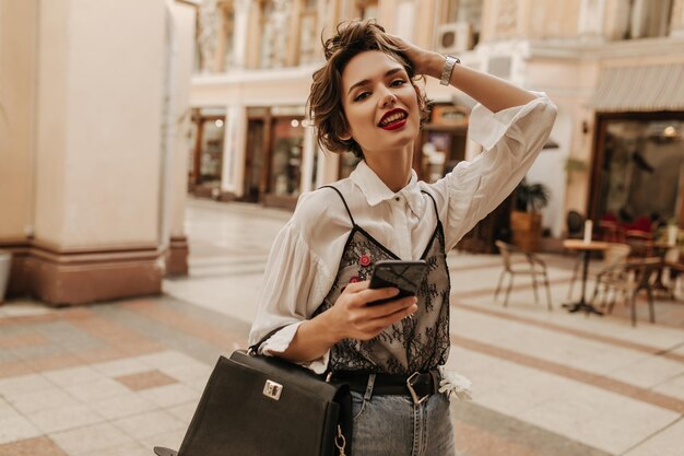 Trendy girl with red lips and short hair in shirt with black lace looking into camera in city Woman holding handbag and phone at street