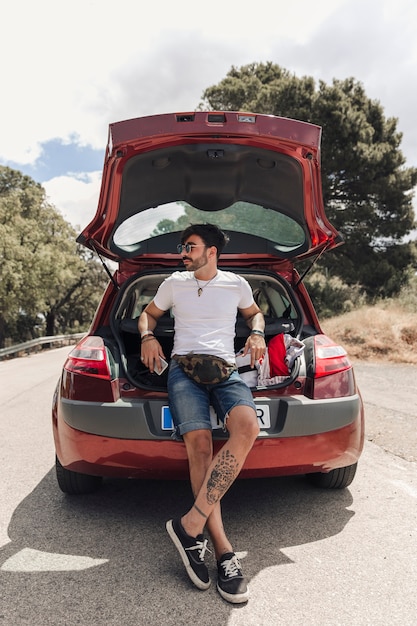 Free photo trendy handsome young man with tattoo leaning on car trunk