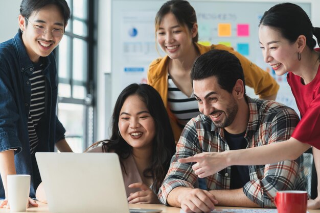 Trendy hipster asian creative friend smiling while sitting at casual meeting with group of friends coworker are working in new success project while using laptop computers digital