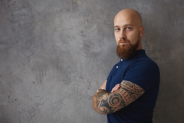 Free Photo trendy looking macho man with muscular tattooed arms and trimmed thick beard posing indoors with arms folded, being displeased or disappointed, pursuing lips, demonstrating negative attitude