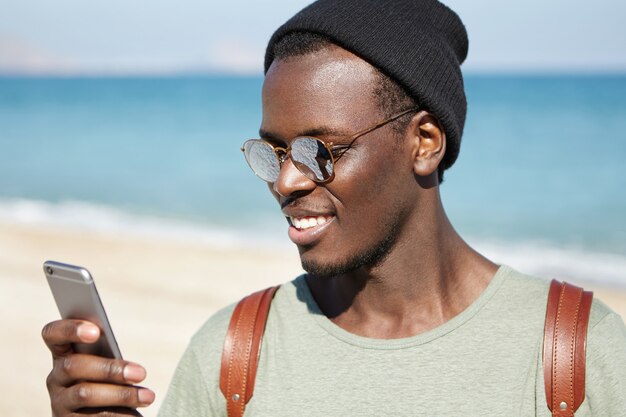 Trendy looking young dark-skinned hipster in mirrored lens shades and headwear reading message or browsing newsfeed via social networks, liking posts and leaving comments while resting on beach