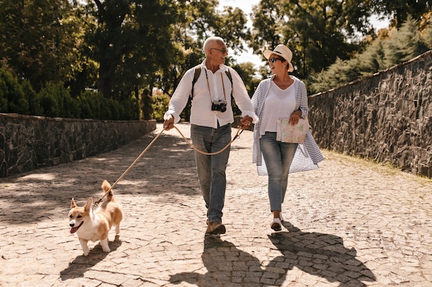 Free photo trendy woman in hat and blue blouse walking and looking grey haired man in white long sleeve shirt with camera and corgi in park.