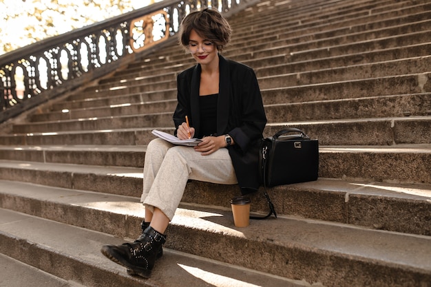 Trendy woman in light pants, black jacket and boots sits on stairs outside. Short-haired lady in glasses writing outdoors.