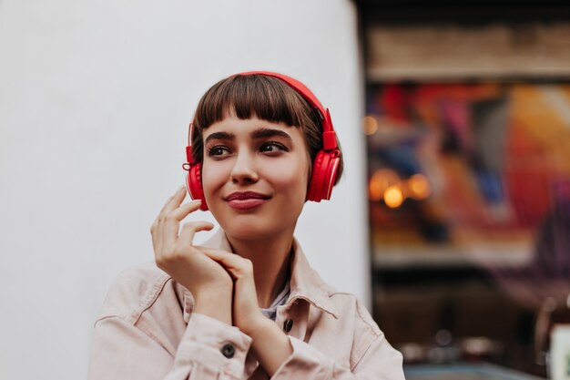 Trendy women in beige denim jacket and right headphones smiling outside