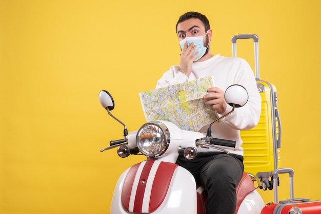 Trip concept with surprised guy in medical mask sitting on motorcycle with yellow suitcase on it and holding map on yellow