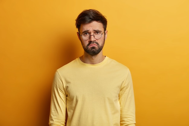 Free Photo troubled displeased bearded man frowns face, feels sad, distressed and upset, being bored sitting on quarantine, unhappy to miss good chance, dressed casually, isolated over yellow wall.