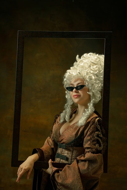 Free photo trying on eyewear. portrait of medieval young woman in vintage clothing with wooden frame on dark background. female model as a duchess, royal person. concept of comparison of eras, fashion, beauty.