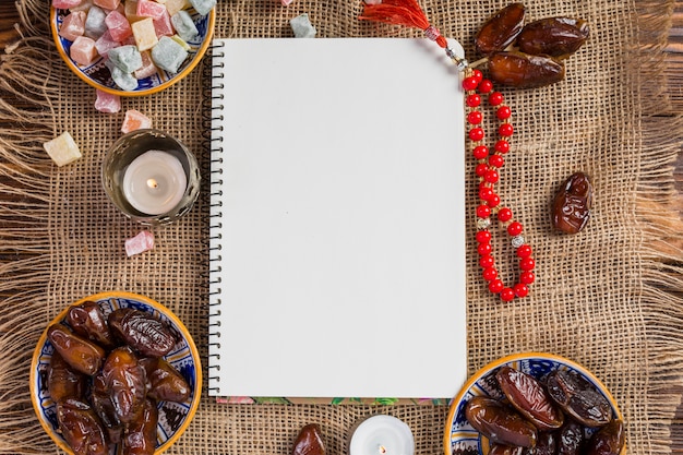 Free Photo turkish bowl of juicy dates; delight lukum with blank white page and red holy beads on jute cloth