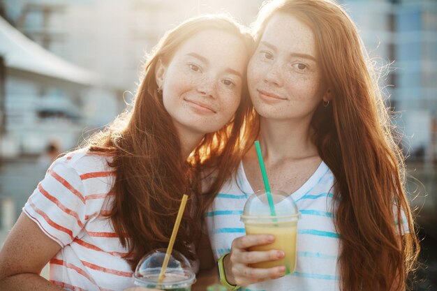 Twin ginger women drinking lemonade looking at camera smiling spending their summer vacation in a city park Warm weather and warm feelings Friendship concept