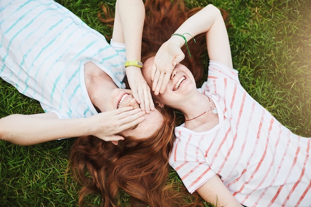 Free photo twin sisters closing their eyes from sun, laying on ground on a summer day.