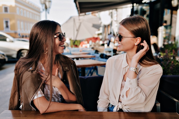 Two adorable smiling ladis in sunglasses sitting and happily talking with friend on summer openair terrace.